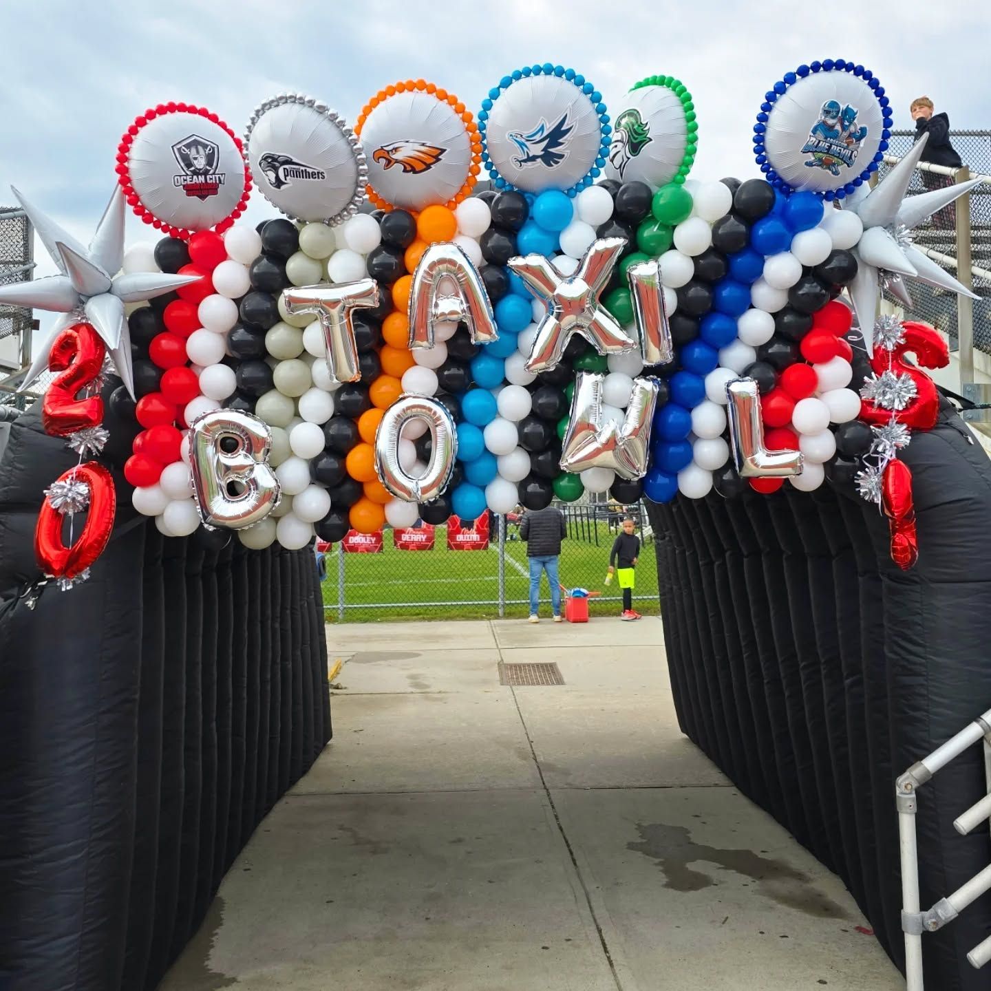 Balloon arch with silver stars and sports logos over a walkway at an outdoor event