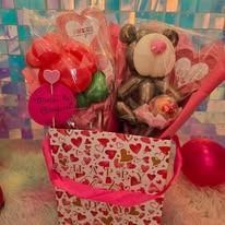 Gift basket with heart-patterned box, pink and red balloons, teddy bear, and candy on a pastel backdrop
