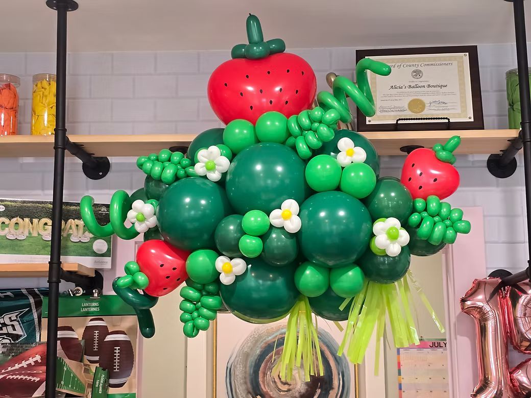 Green and red fruit-shaped balloon bouquet with flowers, displayed indoors on a shelf.