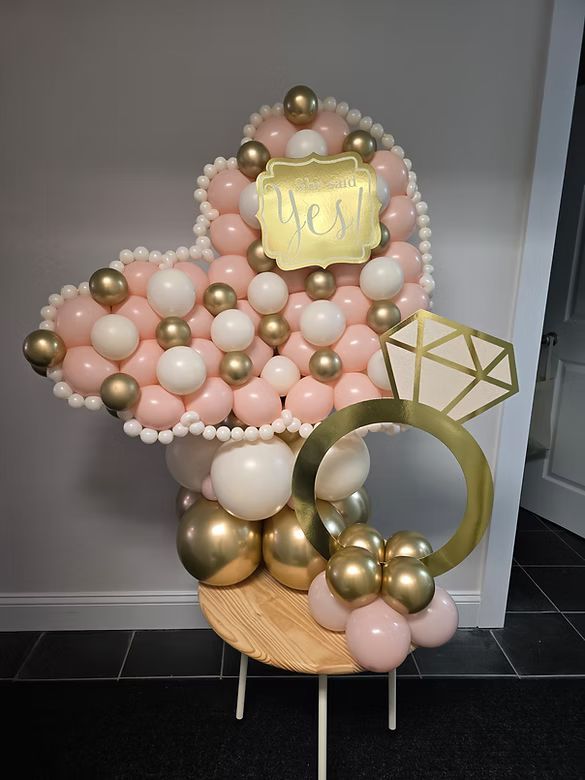 Pastel pink and gold balloon display with a gold “New” sign on a wooden stand.