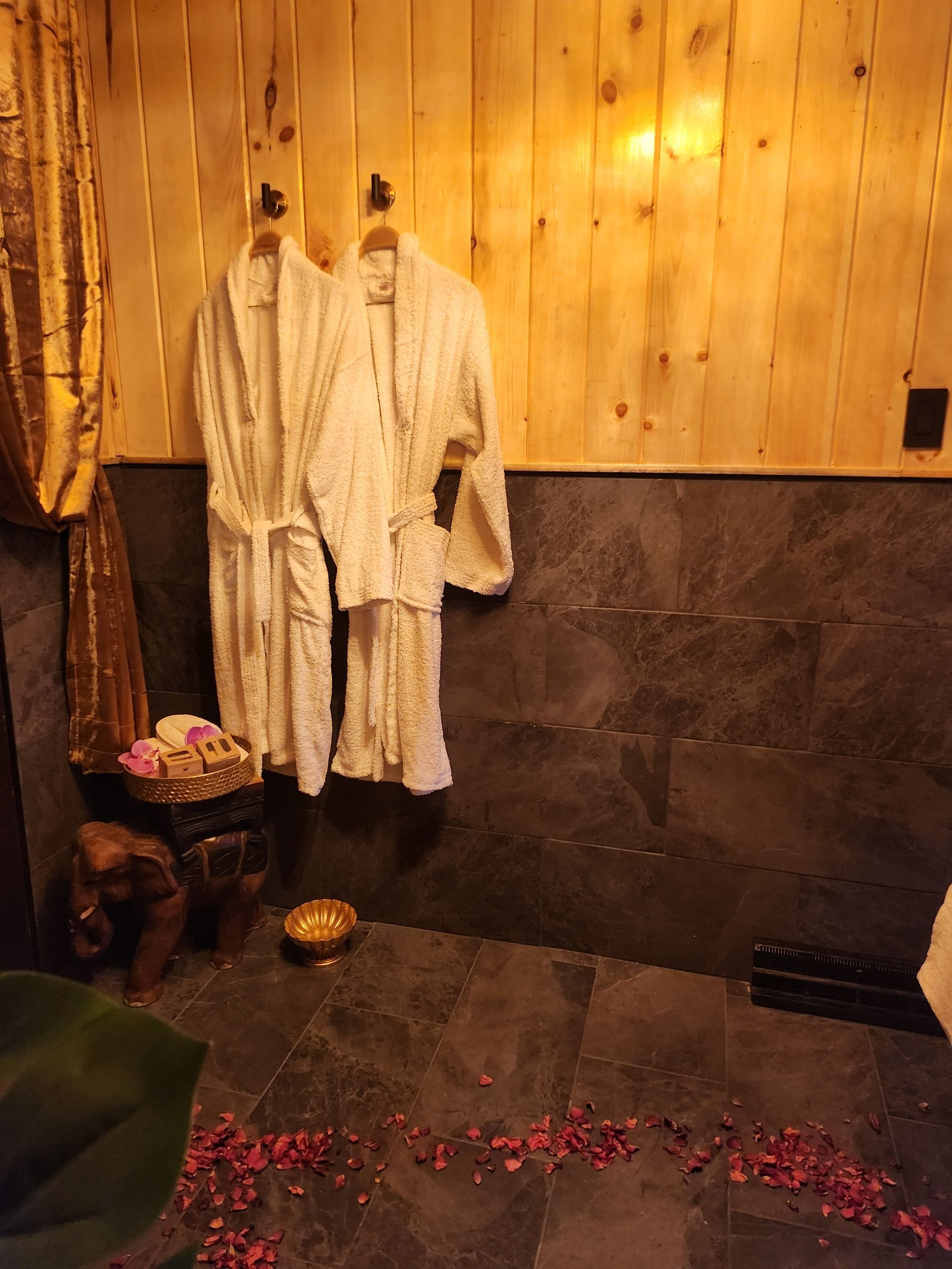Two white robes hang on a wooden wall above a dark tiled area. Rose petals are scattered on the floor.