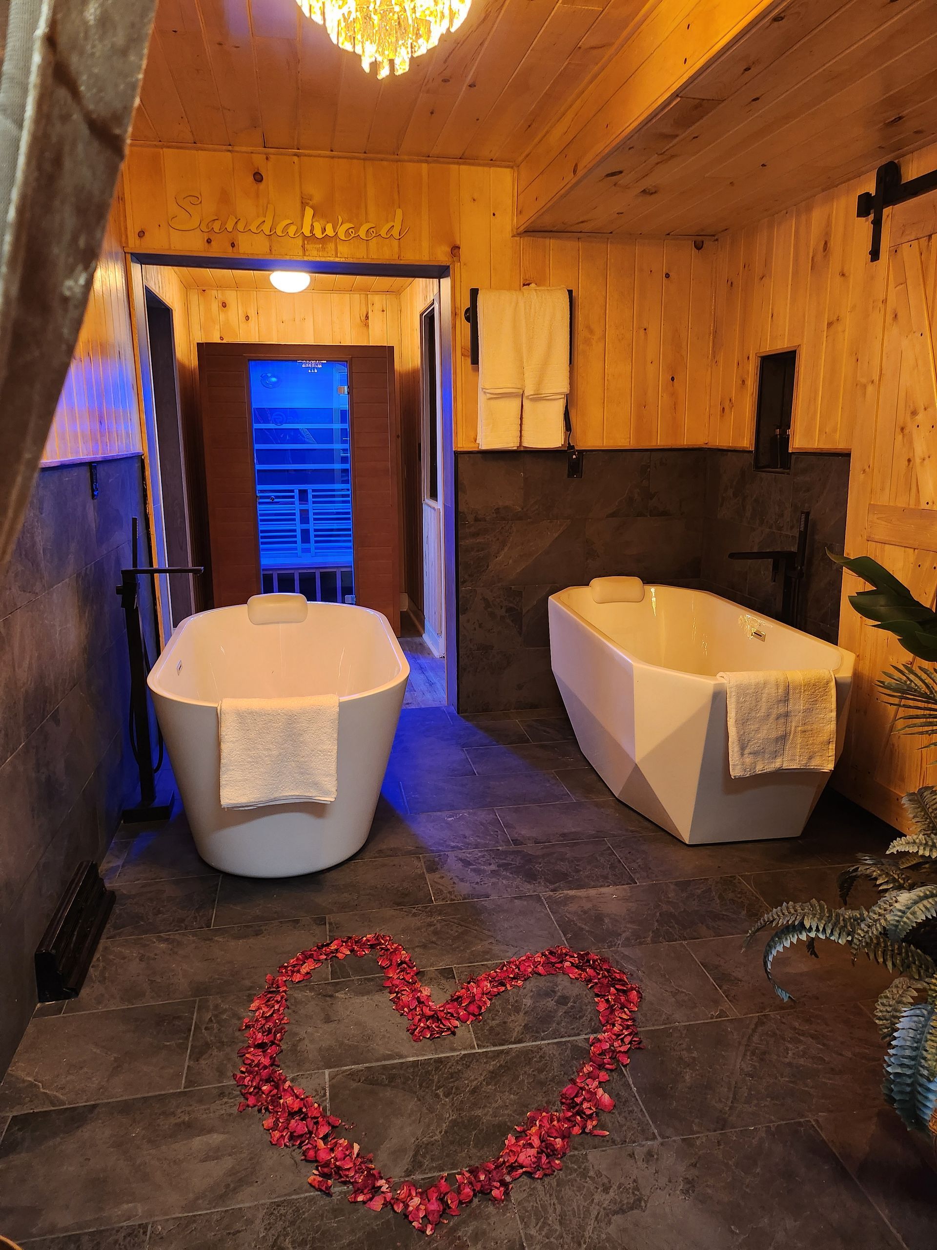 Two white bathtubs in a dimly lit bathroom, with a heart of rose petals on the floor. Wooden walls and a doorway are also visible.