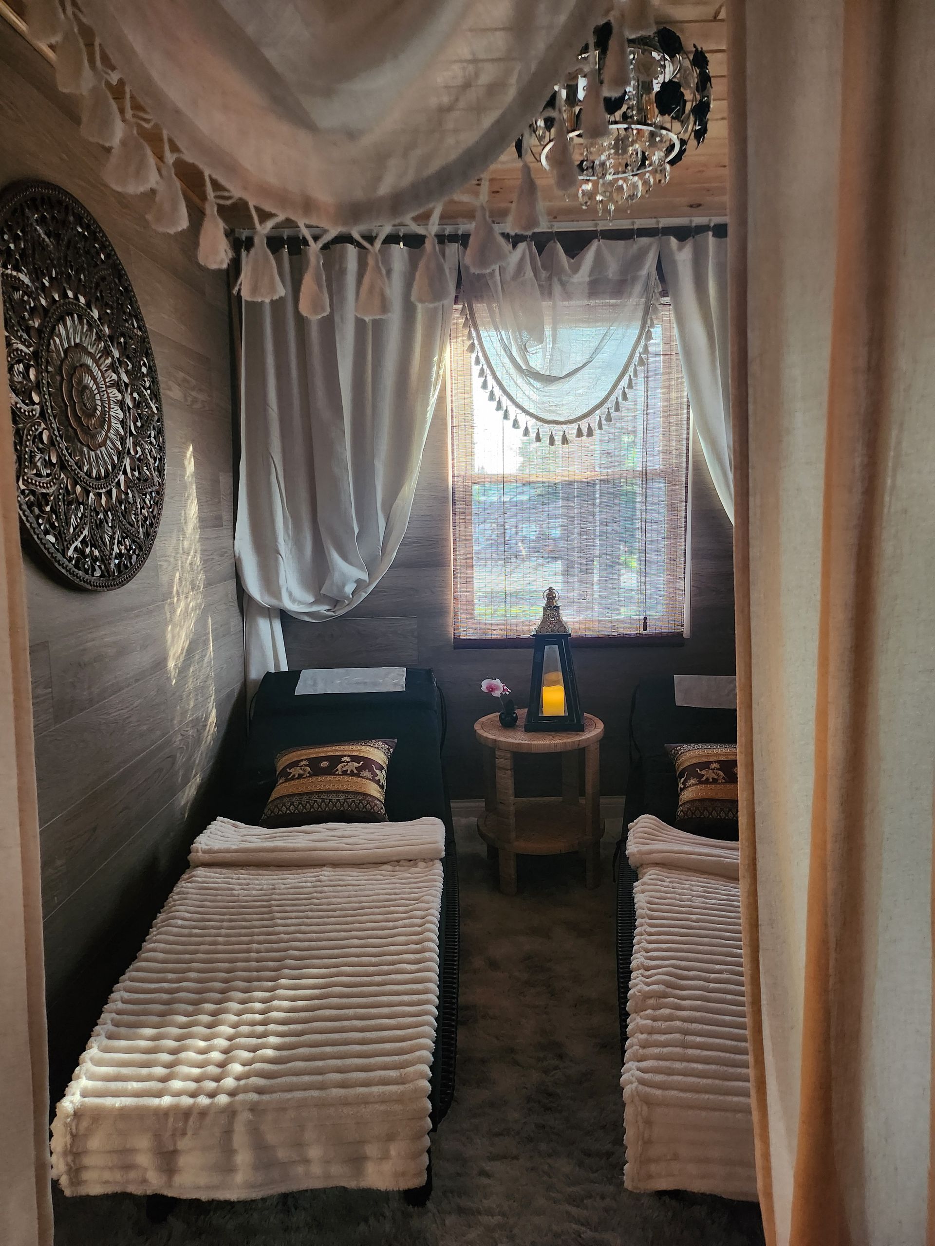 A cozy massage room with two beds, a window, and a decorative wall piece. Soft lighting and draped curtains create a relaxing atmosphere.