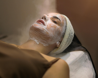 Woman with facial cream lying on a spa treatment bed, eyes closed and wrapped in a towel.