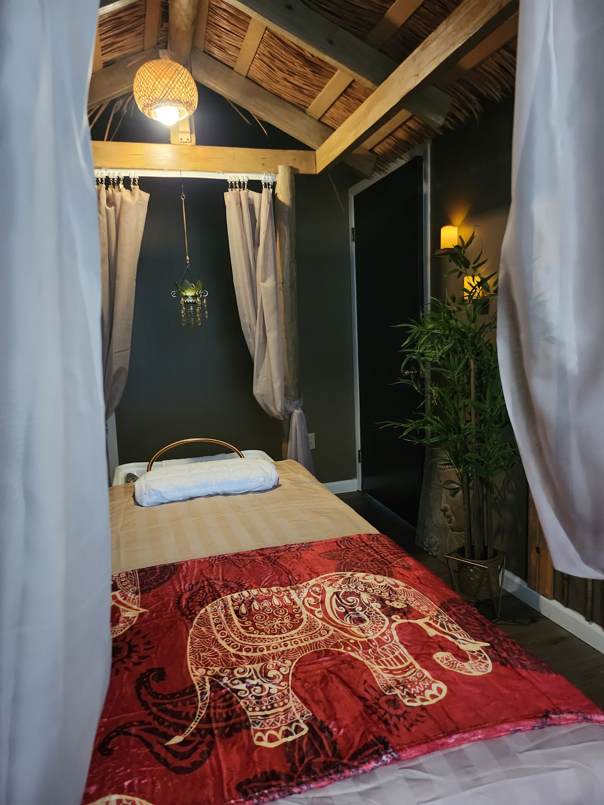 Massage room with red elephant blanket, soft lighting, white curtains, and a decorative bamboo plant.