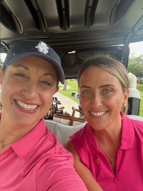 Two smiling golfers in pink shirts sitting in a golf cart