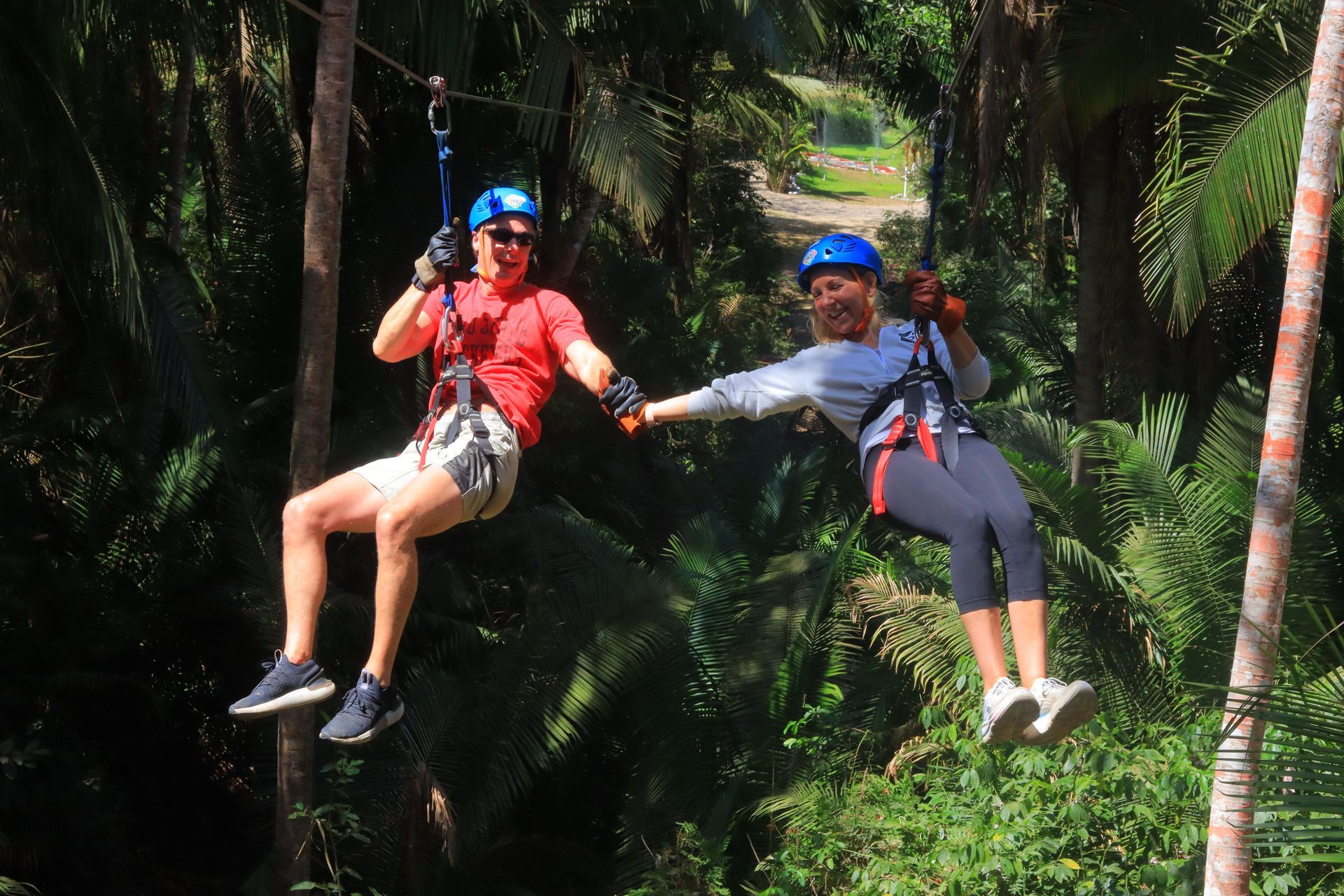 Two people ziplining through a lush green forest, wearing helmets and harnesses.