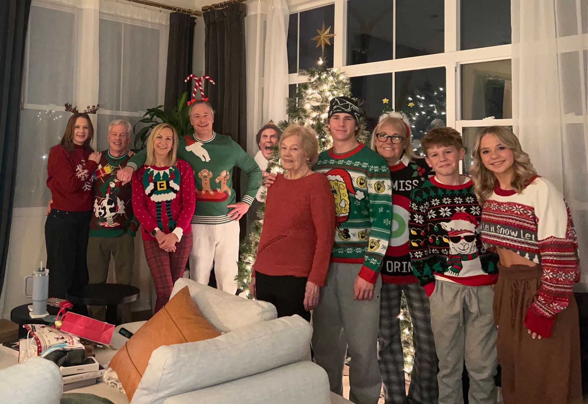 Eight people in festive sweaters posing in a living room decorated with a lit Christmas tree.