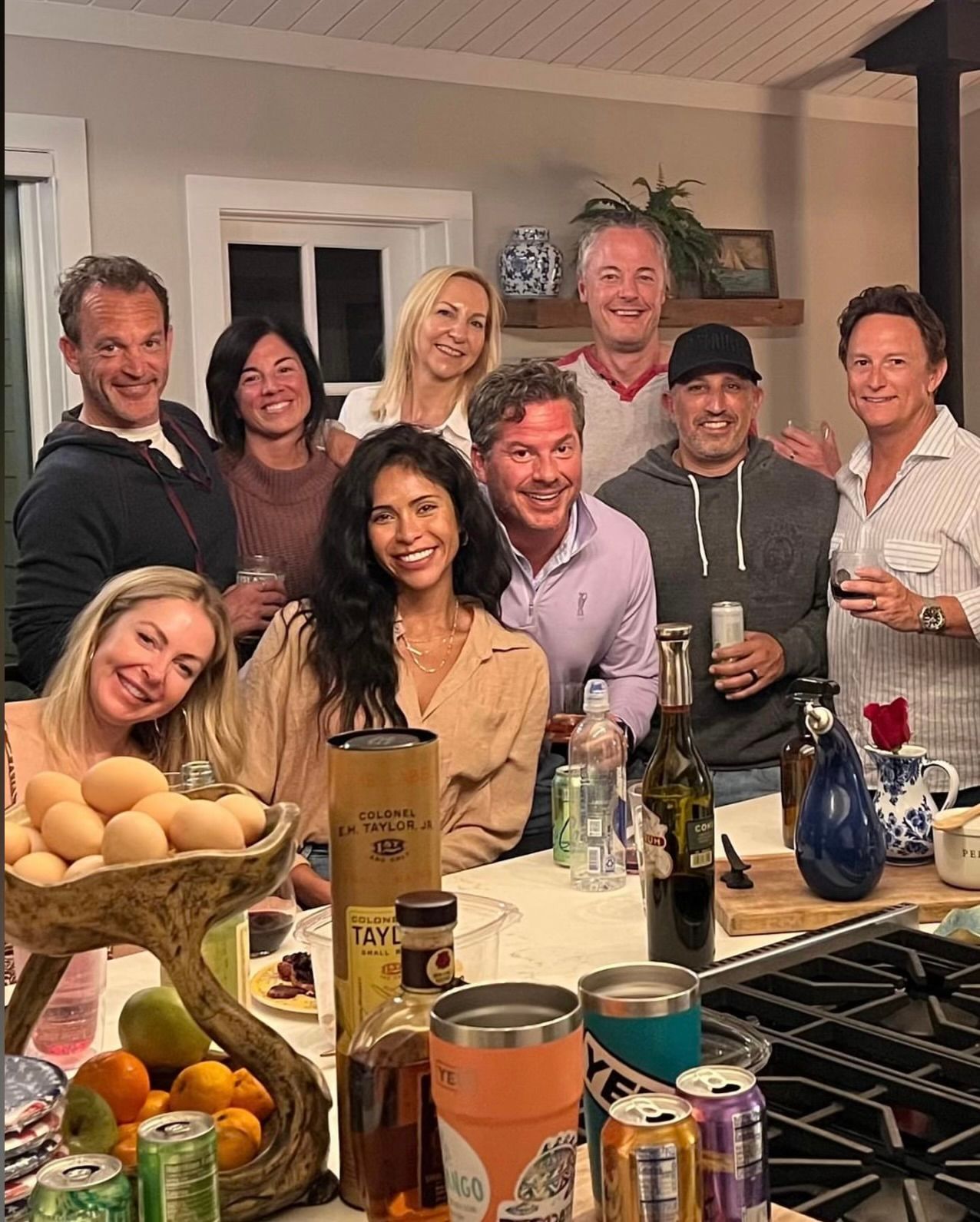 Group of friends smiling around a kitchen counter with drinks, snacks, and fruit during a casual gathering