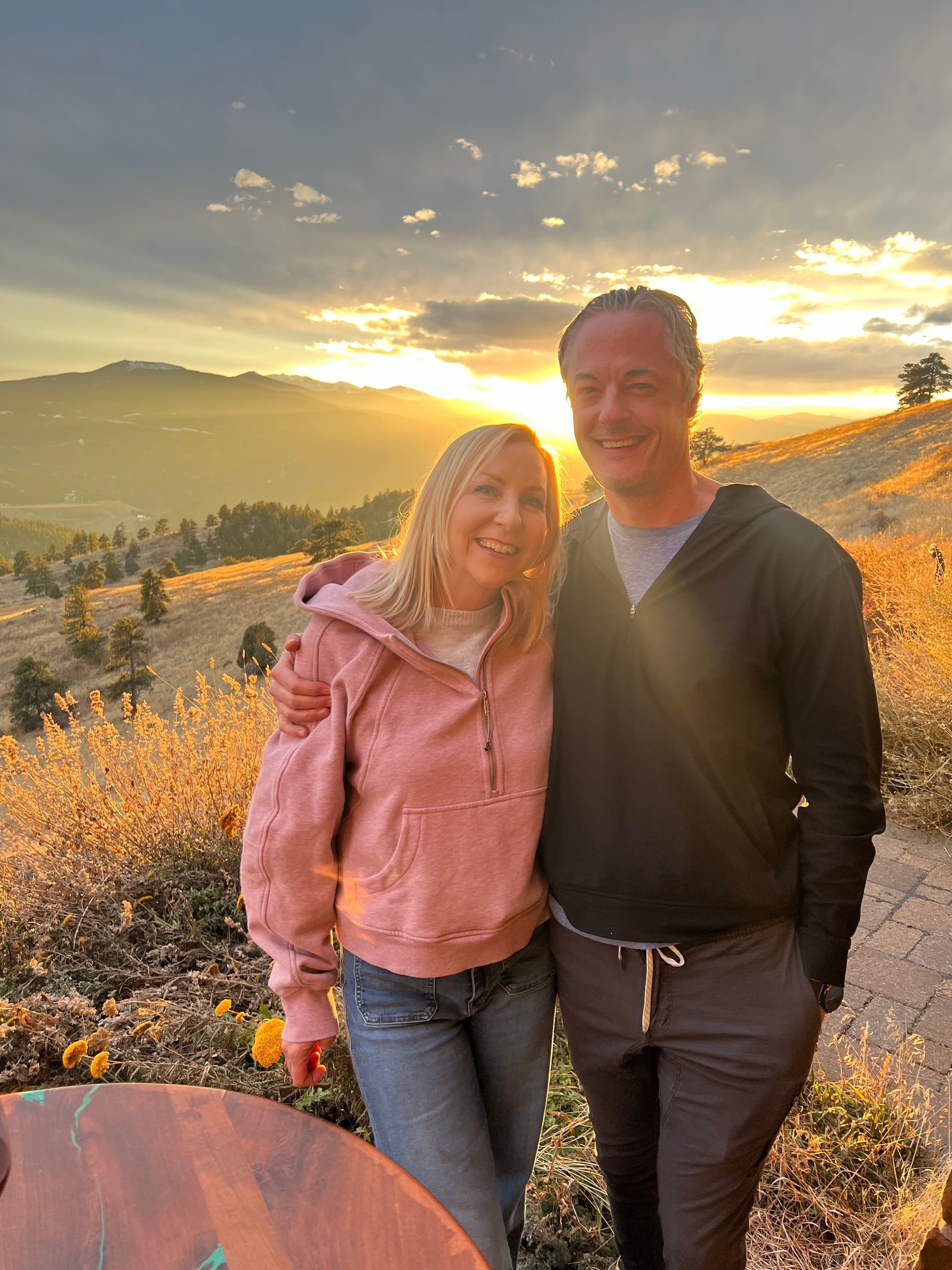 Two people stand together in a field at sunset, with golden light and hills in the background.