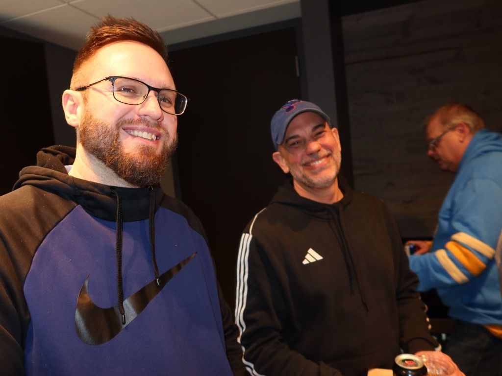Three smiling people indoors, with two facing the camera and one in a blue jacket in the background.
