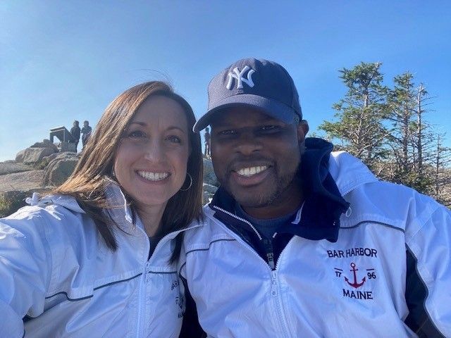 Two smiling people in white jackets and a Yankees cap pose outdoors with blue sky and trees behind them