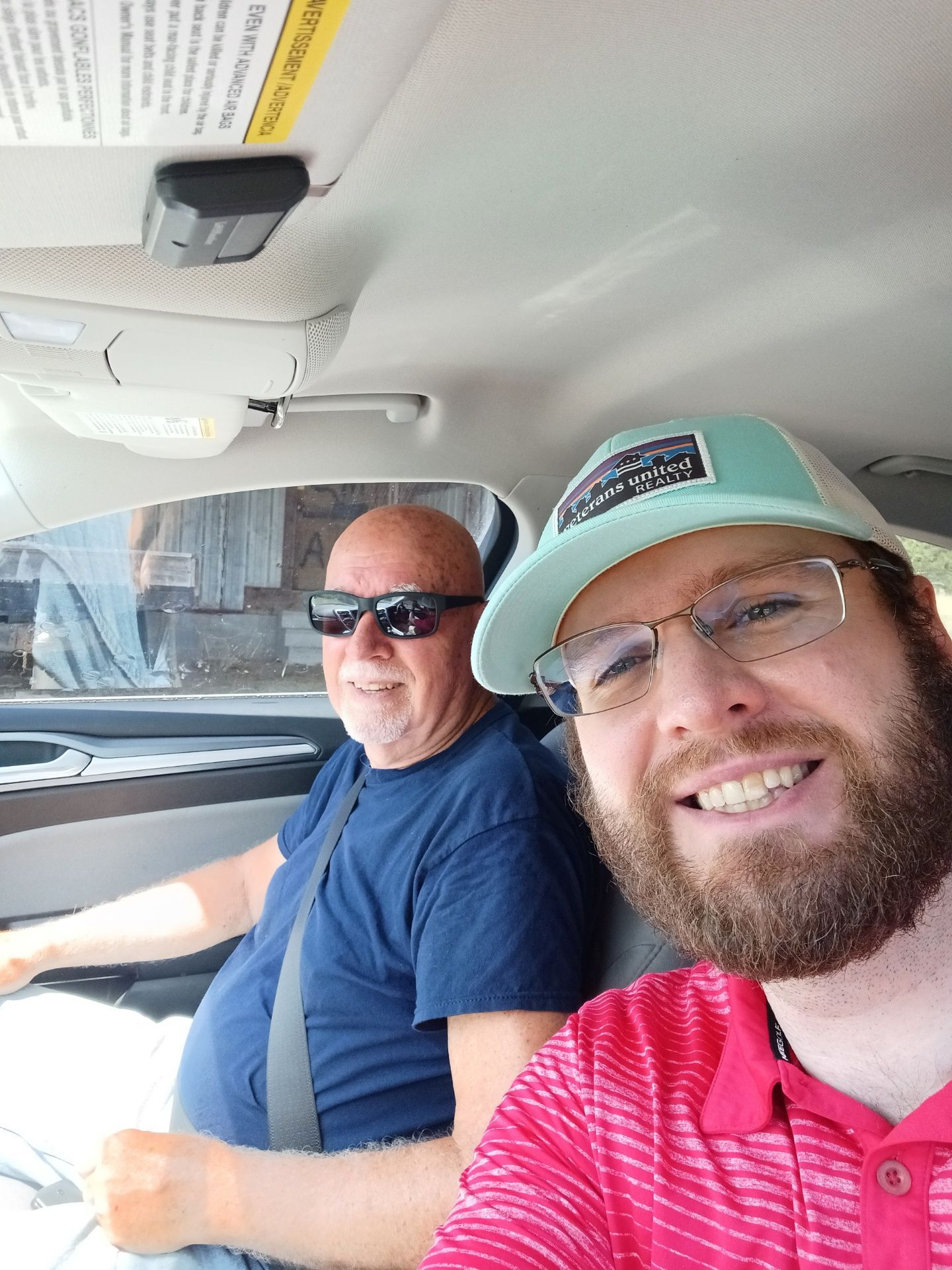 Two men sitting in a car, one smiling in a green cap and red shirt, the other wearing sunglasses and a blue shirt.