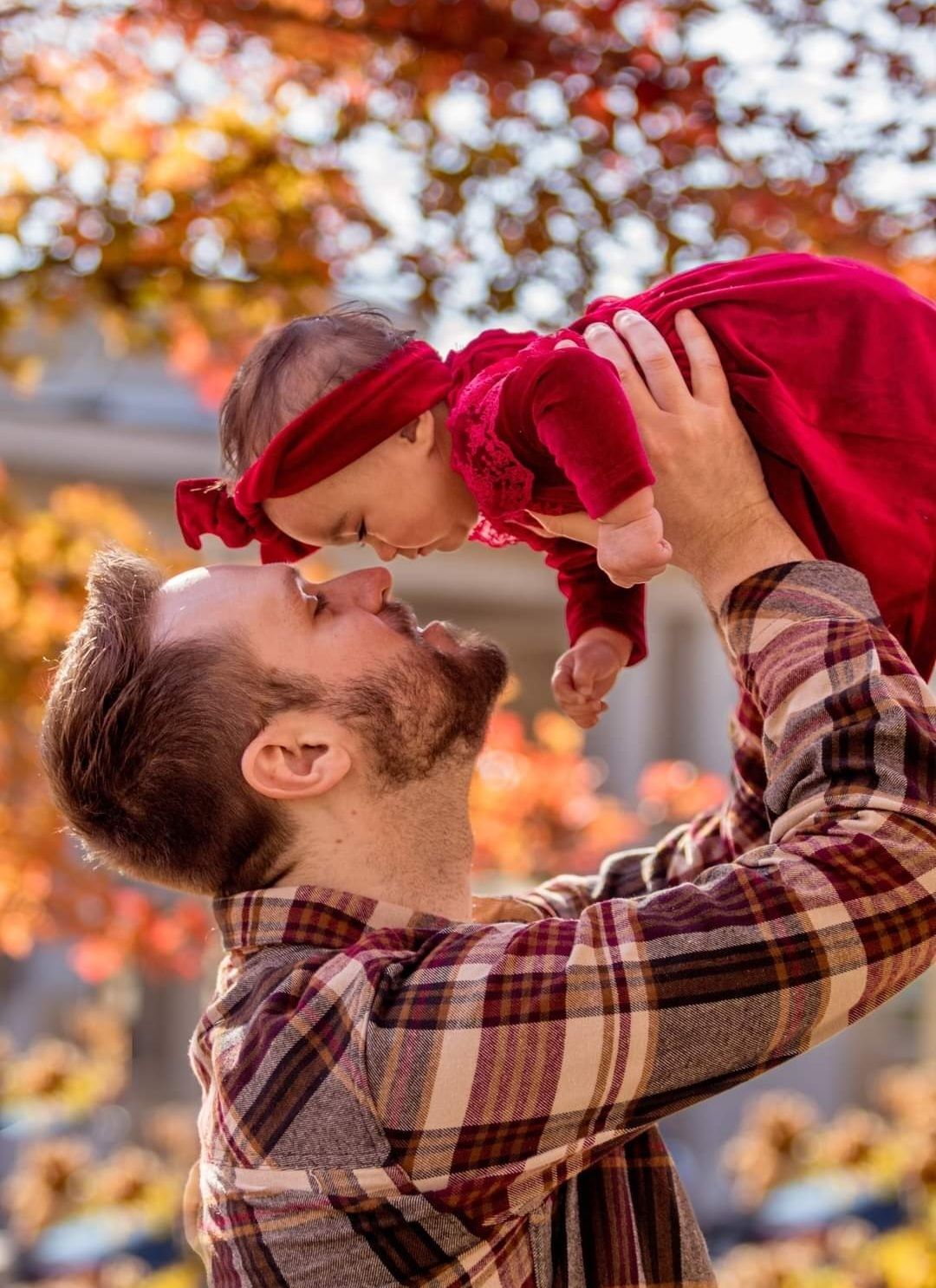 Man lifting a baby in a red outfit outdoors amid autumn leaves