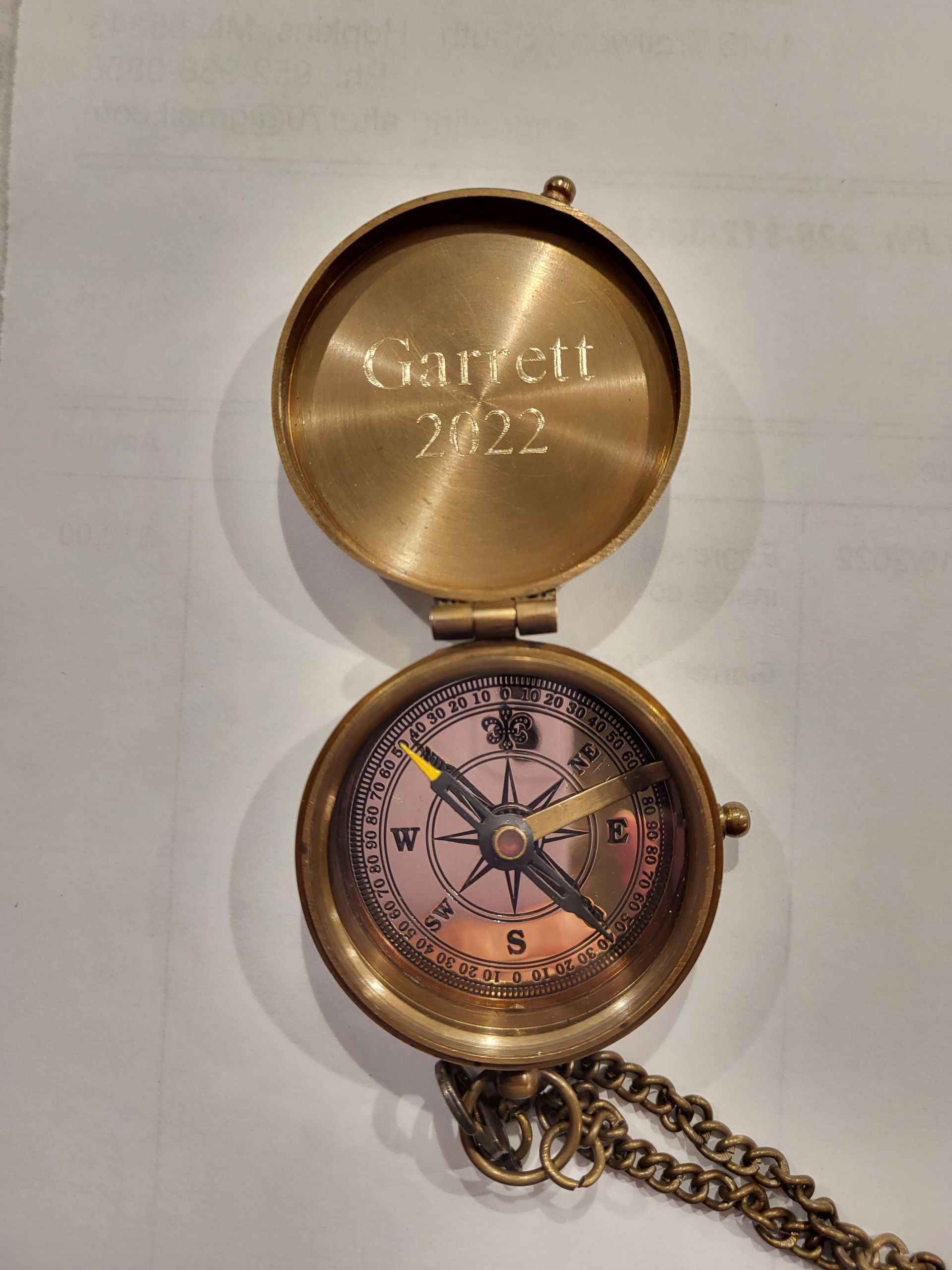 An open, brass-colored pocket compass with an attached chain. The lid is engraved with the name 