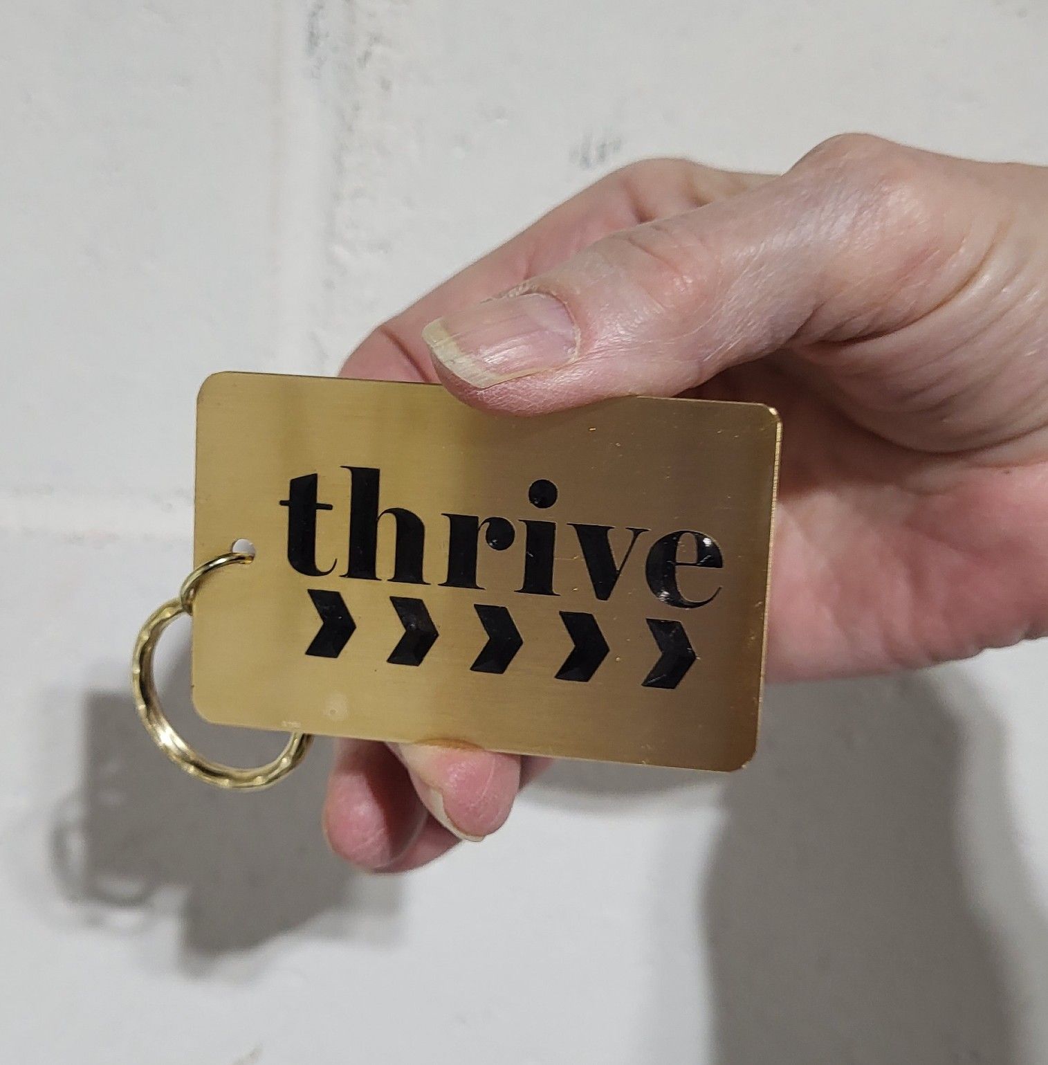 Hand holding a gold card with the word “thrive” and black chevrons, attached to a ring