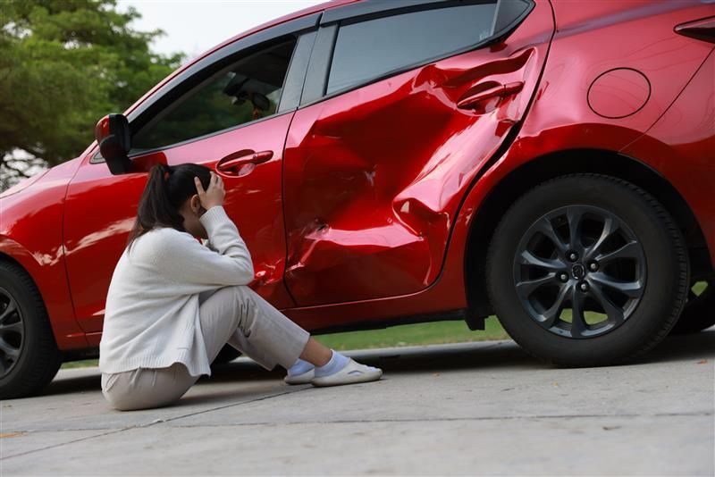 Woman sitting on ground, next to a badly damaged red car; appears distraught.