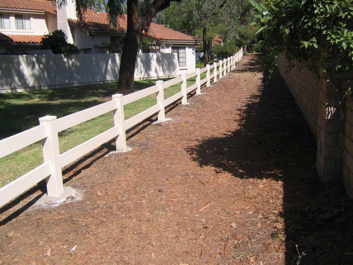 Wooded side path beside a white fence and houses, with sunlight and shadows.
