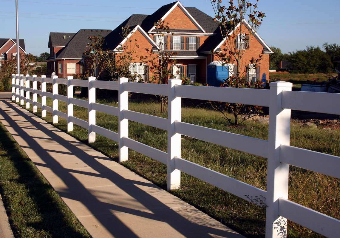 White fence beside a sidewalk in front of brick houses on a sunny suburban street