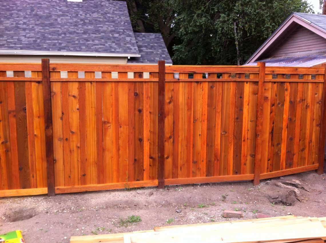 Wooden orange-stained fence with gray posts along a dirt yard, with houses and trees behind it