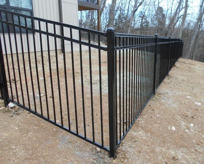 Black metal fence along a gravel path beside a building and wooded slope.