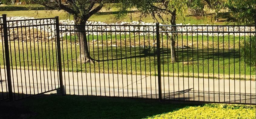 Black metal fence beside a sunlit grassy yard with trees and a paved path in the background