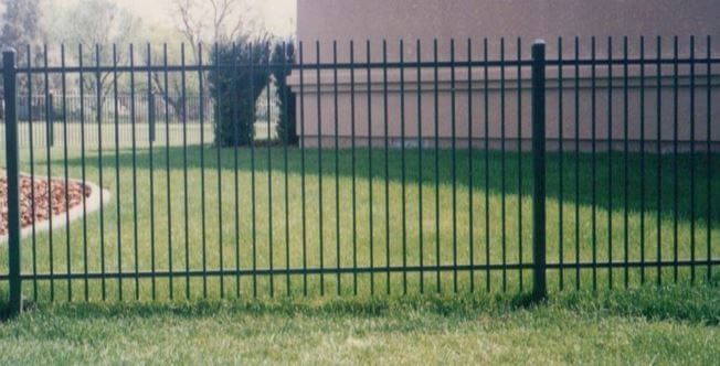 Black metal fence along a grassy yard with trees and a beige wall in the background