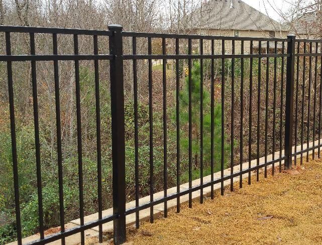 Black metal fence beside a tan sidewalk, with trees and brush in the background