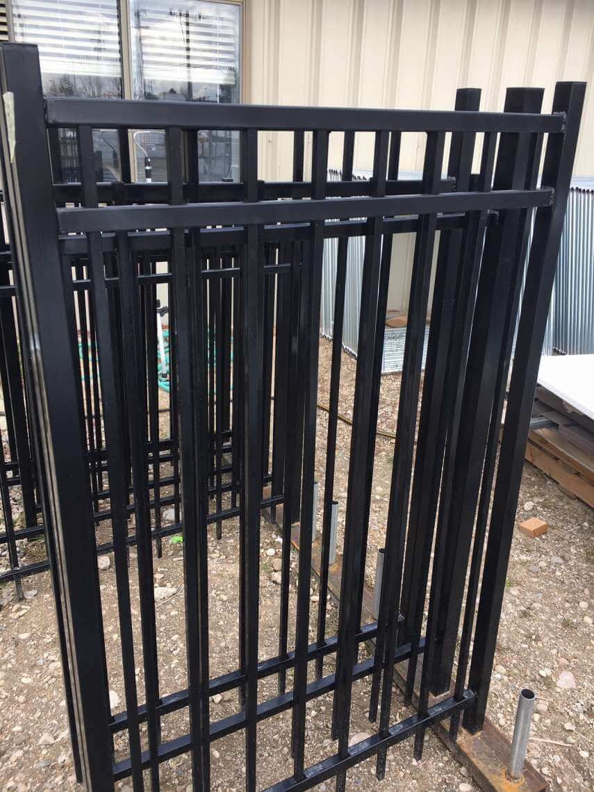 Black metal gate panels leaning outdoors on gravel beside a building and windows