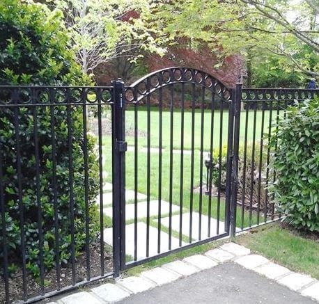 Black wrought-iron garden gate between hedges, opening to a lawn and stone path.
