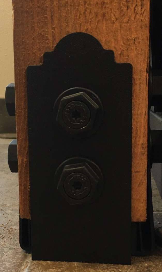 Black metal bracket with two bolts attached to a wooden beam.