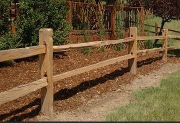 Wooden split-rail fence along a mulch path beside green grass and shrubs