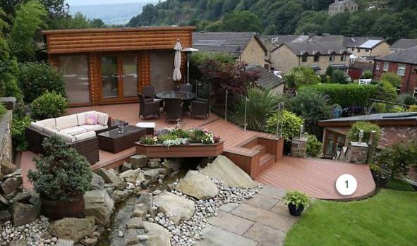 Modern backyard patio with wooden deck, lounge seating, fire pit, pond, and stone garden path under a house hillside.