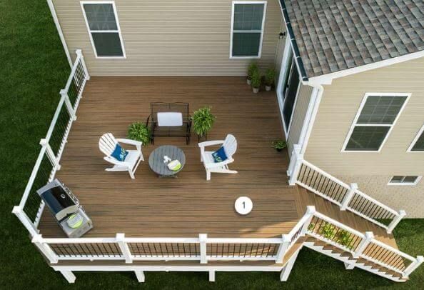 Aerial view of a wooden backyard deck with white railing, outdoor chairs, table, grill, and potted plants.