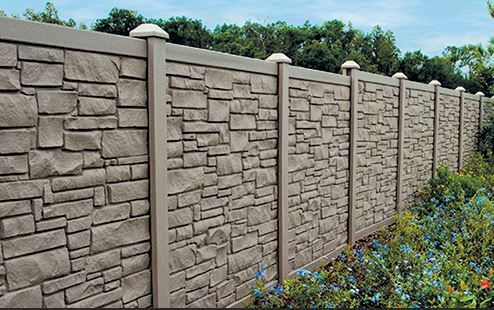 Stone-textured privacy fence with white posts and greenery in front under a blue sky