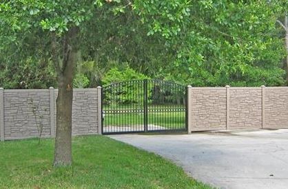 Stone wall with a black metal gate opening to a green yard, beside a tree and driveway.