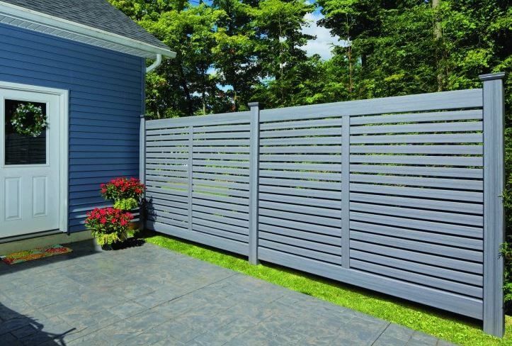 Gray slatted privacy fence beside a blue house with a white door and potted red flowers