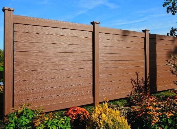 Brown wooden privacy fence with decorative panels beside colorful landscaping under a blue sky