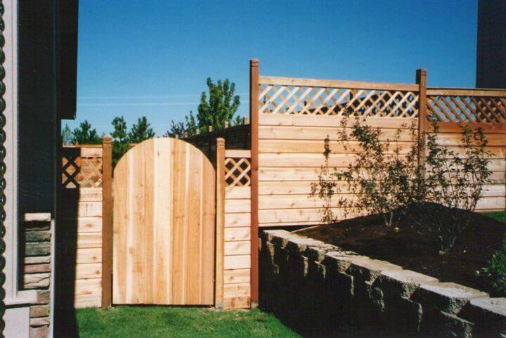 Wooden garden gate between brick pillars and a lattice fence beside a stone retaining wall under a blue sky