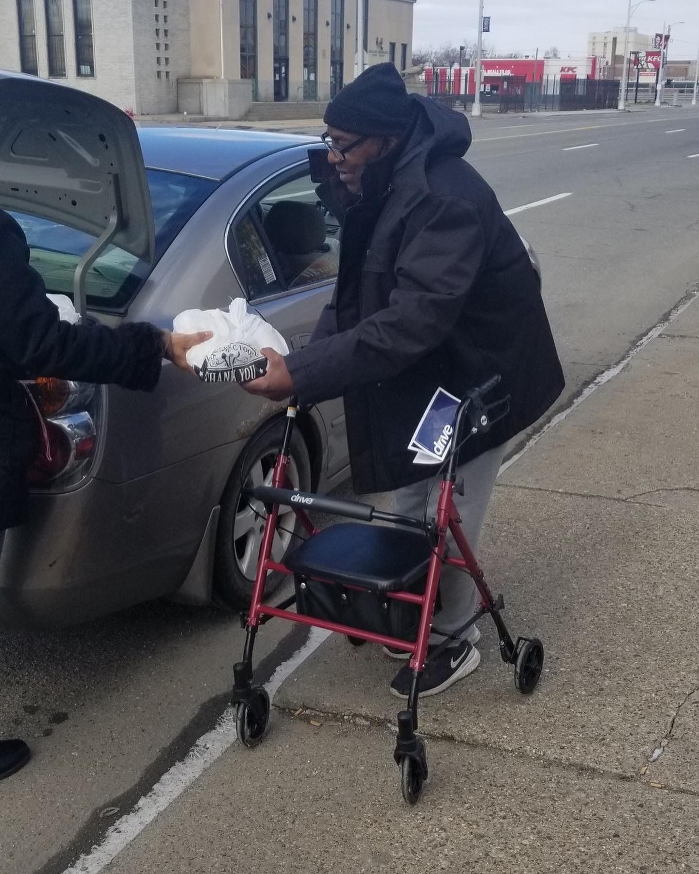 Person with walker receiving a bag from a car on a city sidewalk