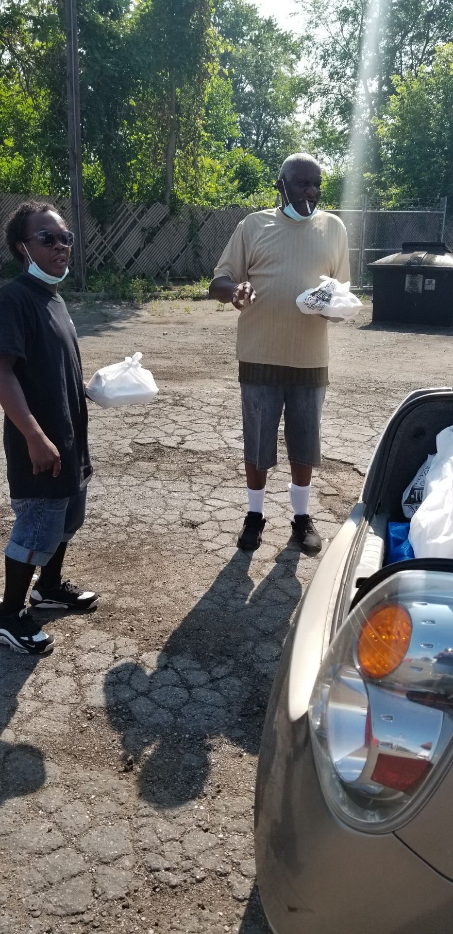 Two people holding white bags near a car on a sunny outdoor path