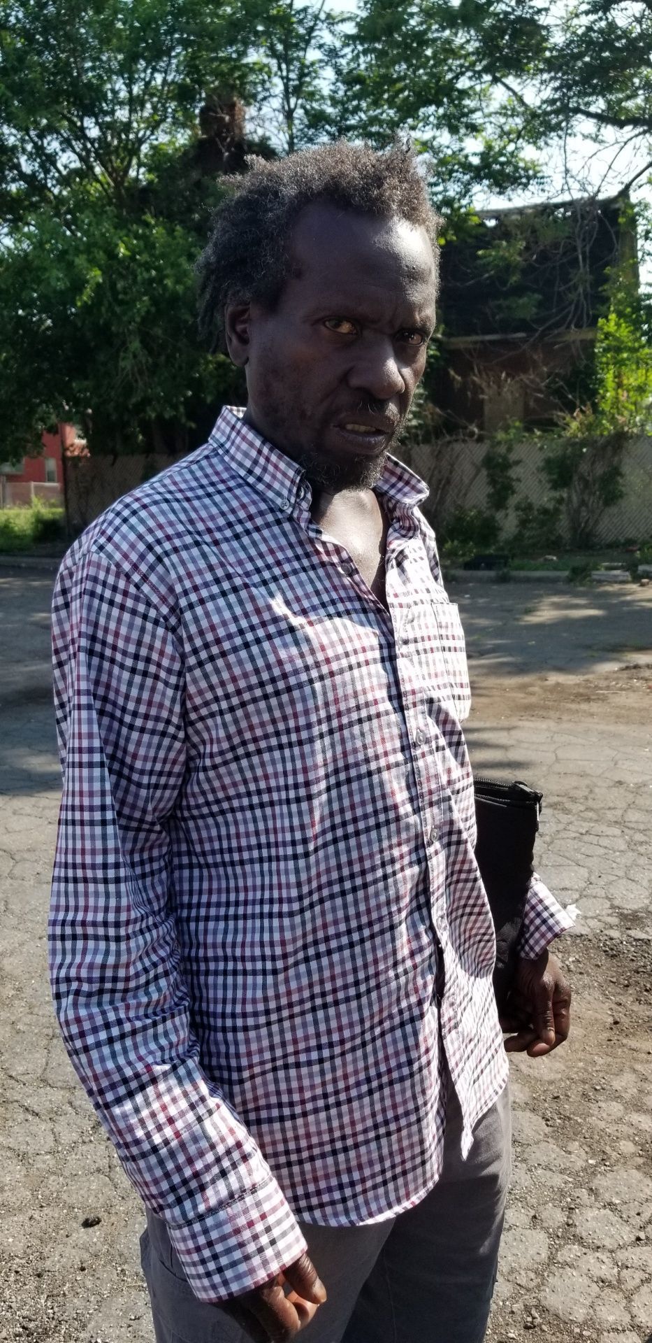 Man standing outdoors in a checkered shirt, holding papers on a sunny street.