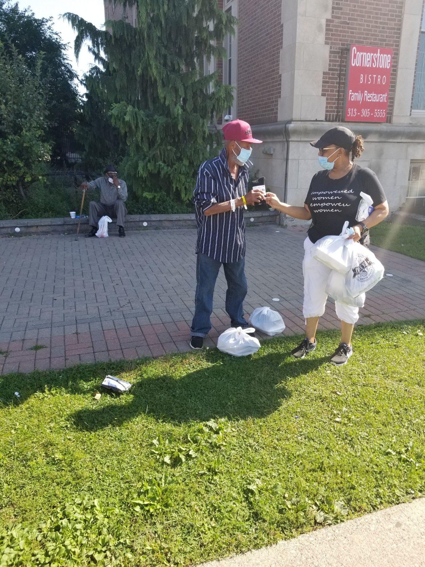 Two people exchanging items on a sunny sidewalk beside a grassy area and building.