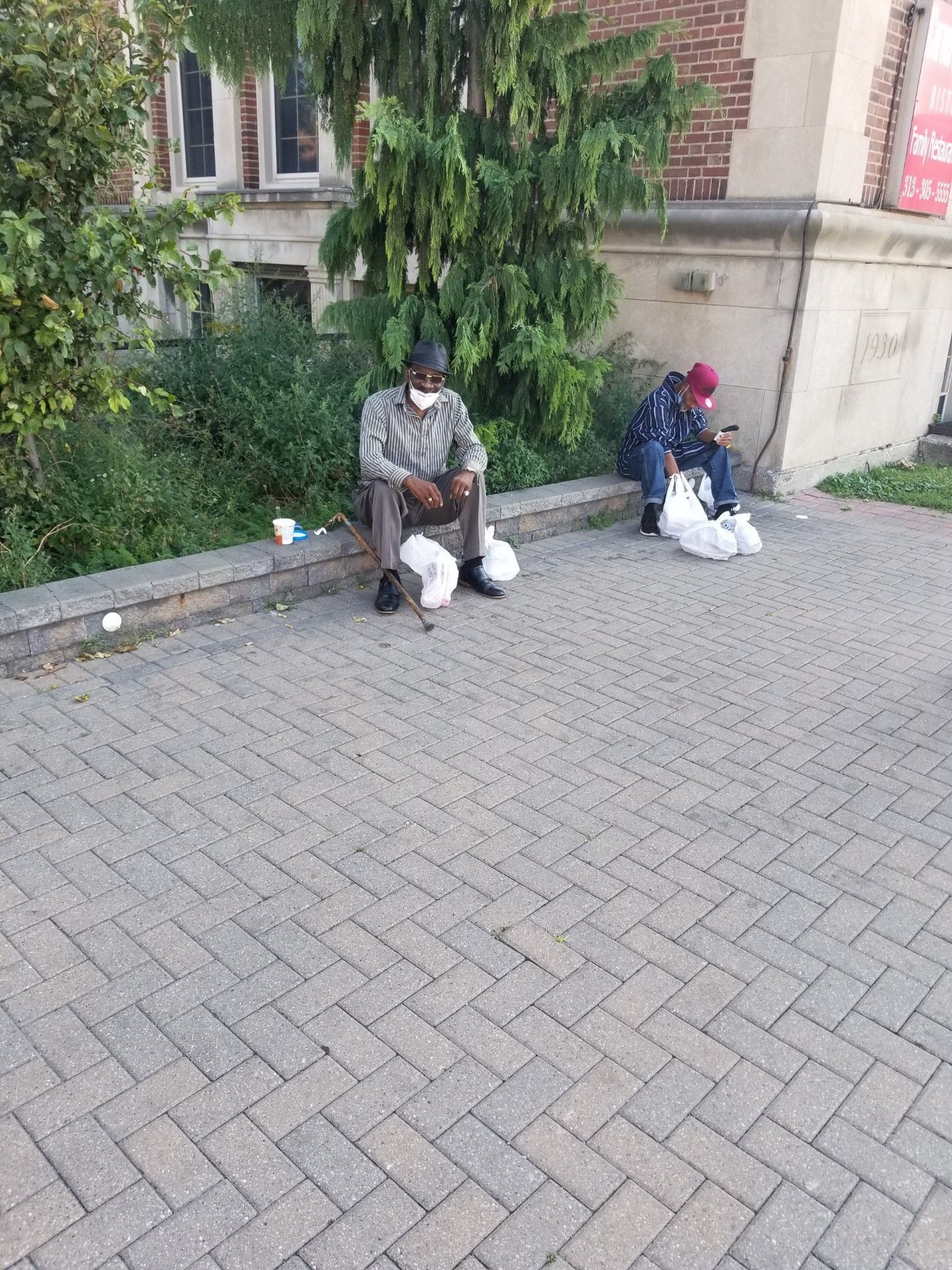 Two people sitting on a curb beside a brick building, each with bags, on a paved street.