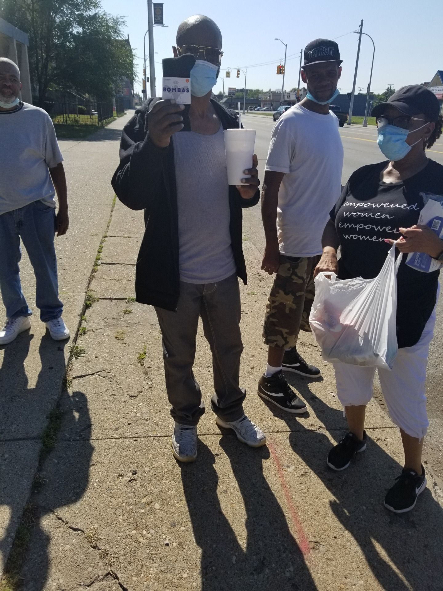Group of masked people standing outdoors on a sunny sidewalk, one holding a sign and another holding a plastic bag.