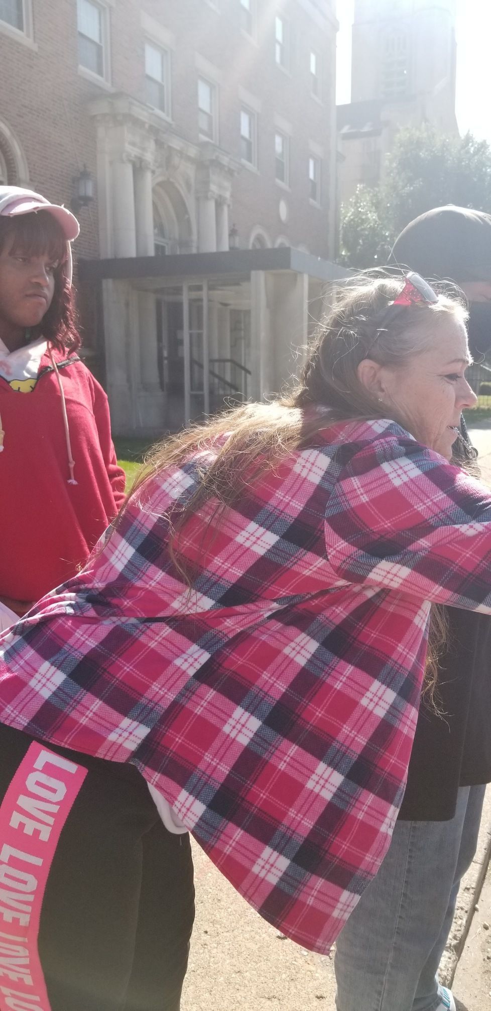 Two people outdoors, one in a red plaid coat leaning forward, near a brick building in bright sunlight