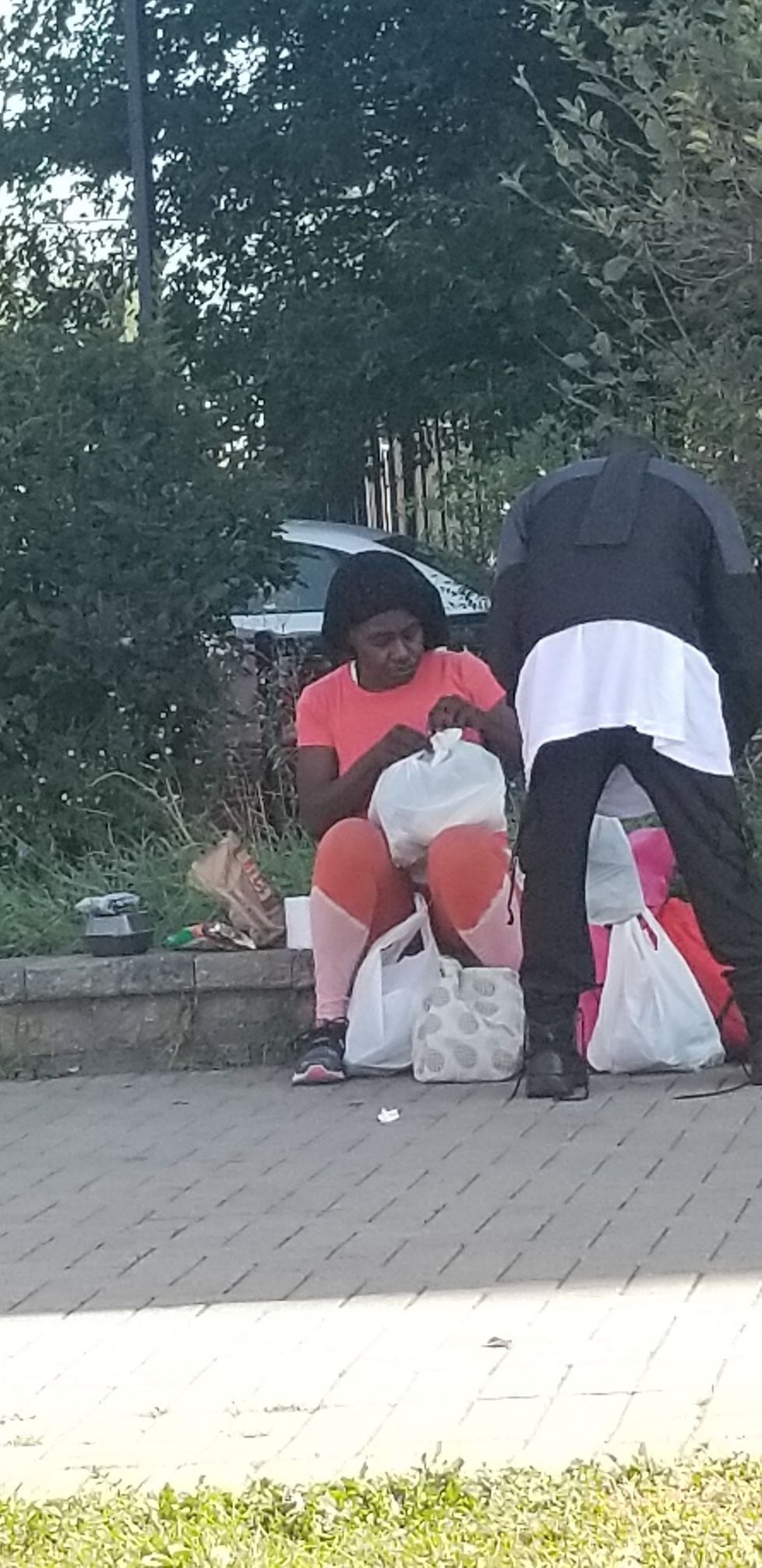Two people sit on bags on a sidewalk near trees, one in a red top and one in dark clothes.