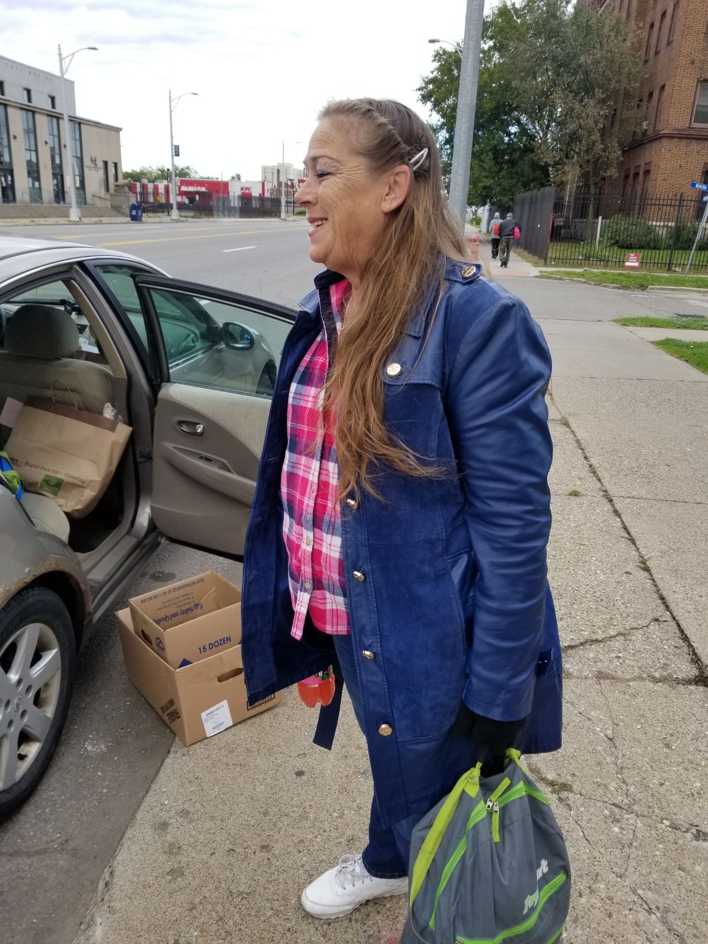 Woman standing beside open car door, holding a green bag and smiling on a sidewalk with boxes nearby.