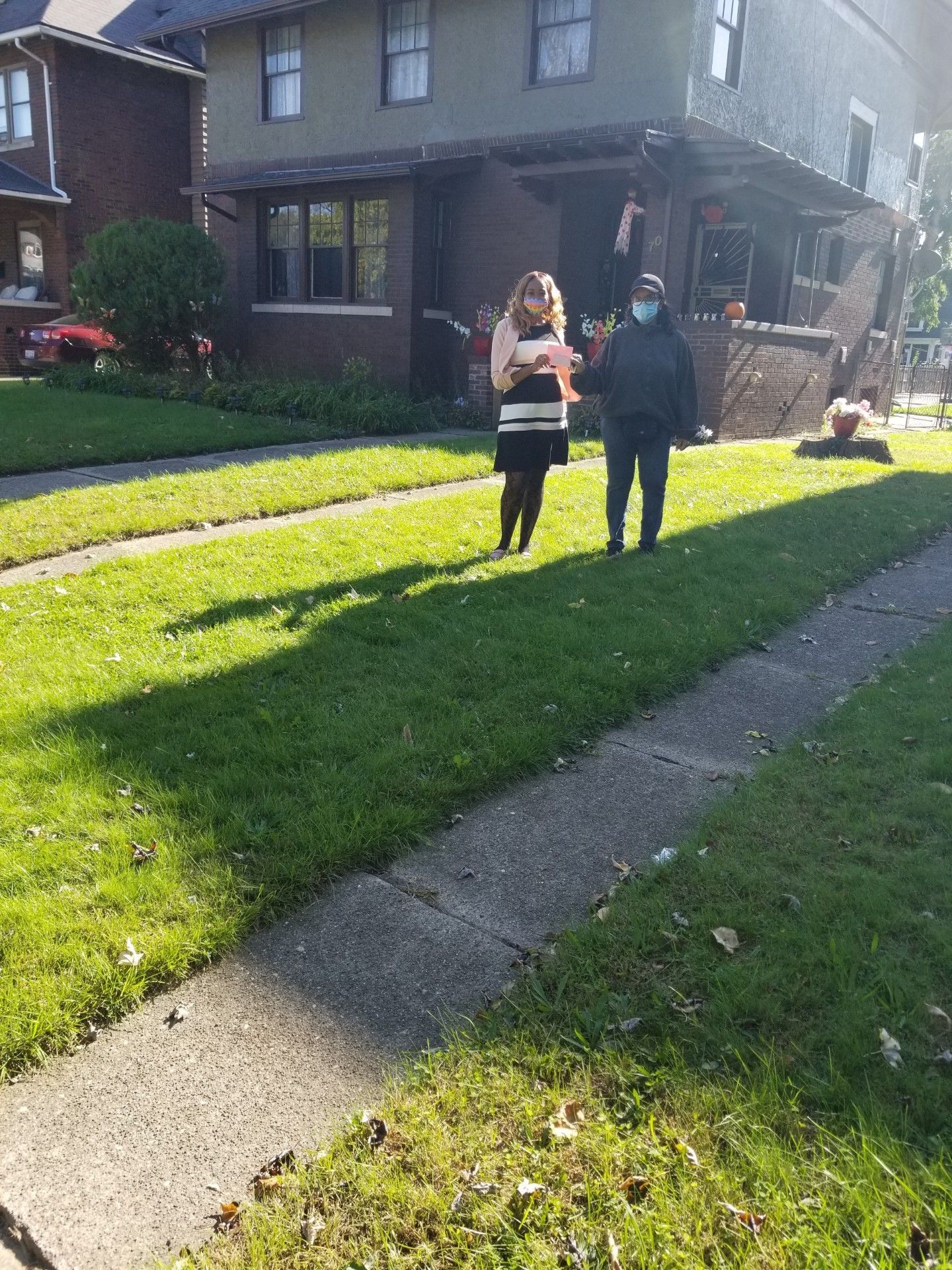 Two people standing on a sunny sidewalk in front of a brick house and green lawn