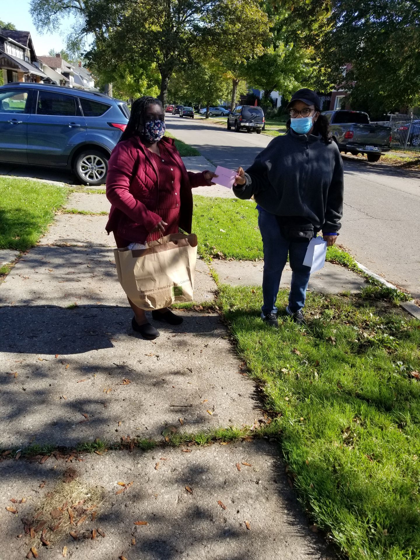Two masked people walking on a sidewalk in a sunny neighborhood, one carrying a paper bag